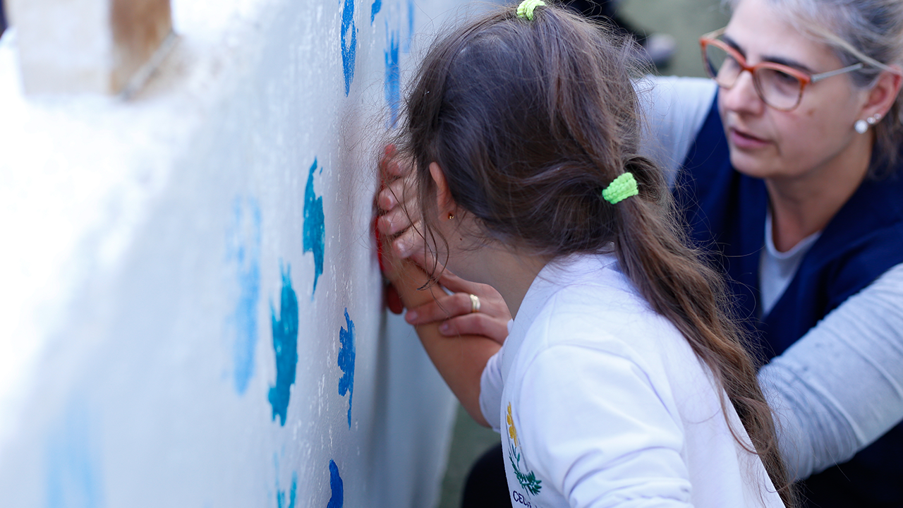 Uma aluna da Escola CEDAE está pintando. Na foto. com a ajuda de uma professora, ela pressiona a mão com tinta na parede para ficar marcado o formato da mão