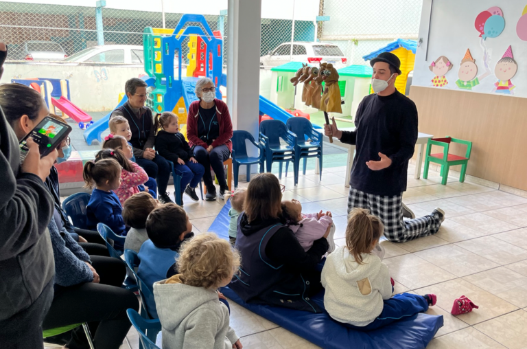 Alunos do CEDAE acompanham sentados de costas para a foto a apresentação de teatro de fantoches. Mais ao fundo, Iury apresenta o teatro de joelhos, ele está com cavalinhos na mão.