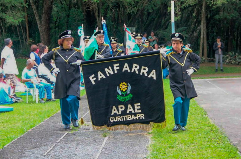 A imagem mostra os alunos que participam da fanfarra segurando bandeiras. Na frente aparecem dois alunos segurando uma bandeira escrito: ‘’Fanfarra Apae Curitiba’’ com o símbolo da instituição no meio.