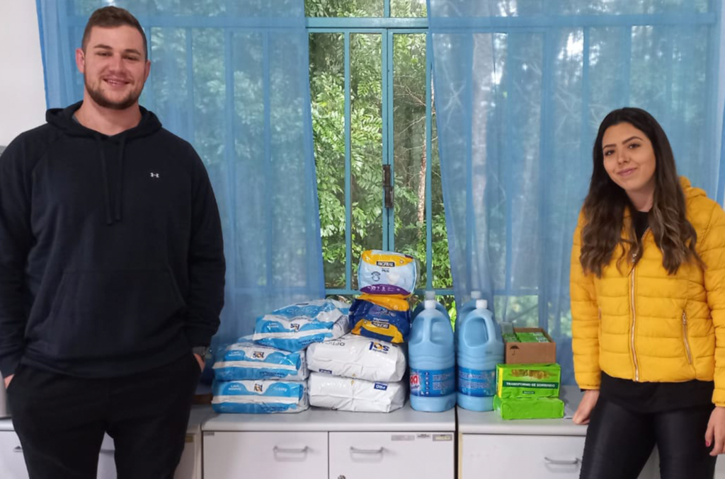 Dois alunos posam para a foto. Um menino está do lado esquerdo e a menina do lado direito. Ao fundo, no centro, os produtos de higiene e limpeza estão sobre um armário