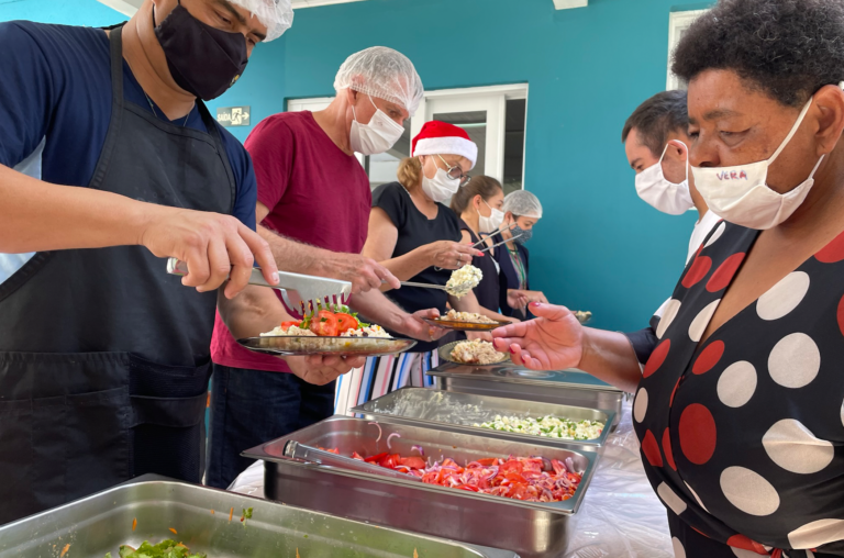 do lado esquerdo cinco pessoas estão servindo pratos de comida para as pessoas com deficiência que estão do lado direito. Ao centro, formas com comidas.
