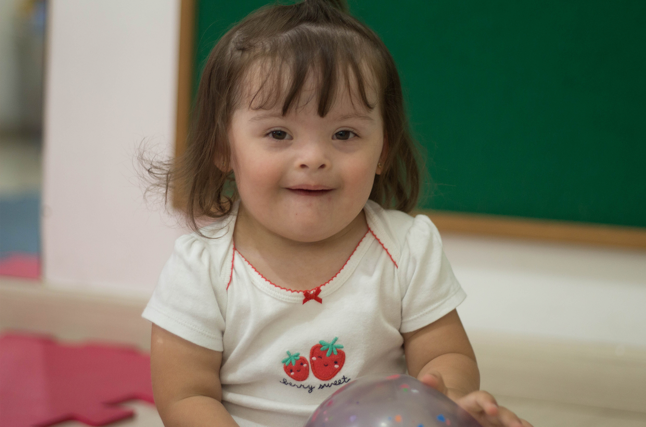 Aluna da escola CEDAE dentro da sala de aula, sentada no chão, com uma camisa branca com o desenho de dois morangos, ela sorri