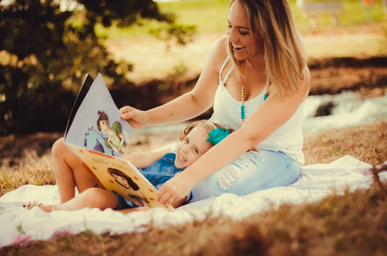 A foto mostra a mãe sentada lendo para o filho no meio do parque