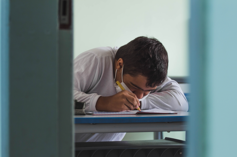Estudante sentado, dentro de uma sala de aula, escrevendo