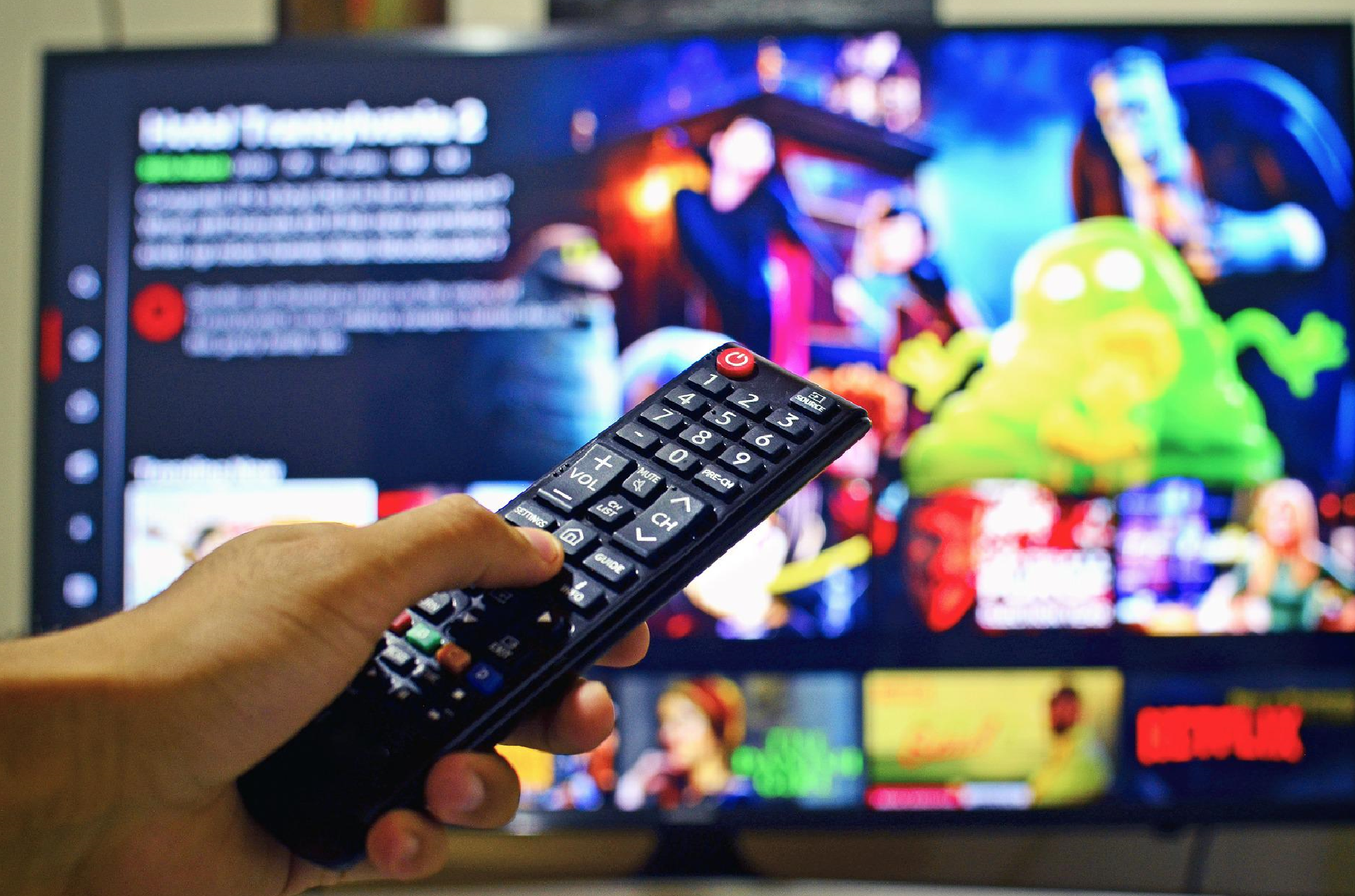 Foto de uma mão segurando um controle remoto, ao fundo, desfocado uma televisão ligada em um desenho animado